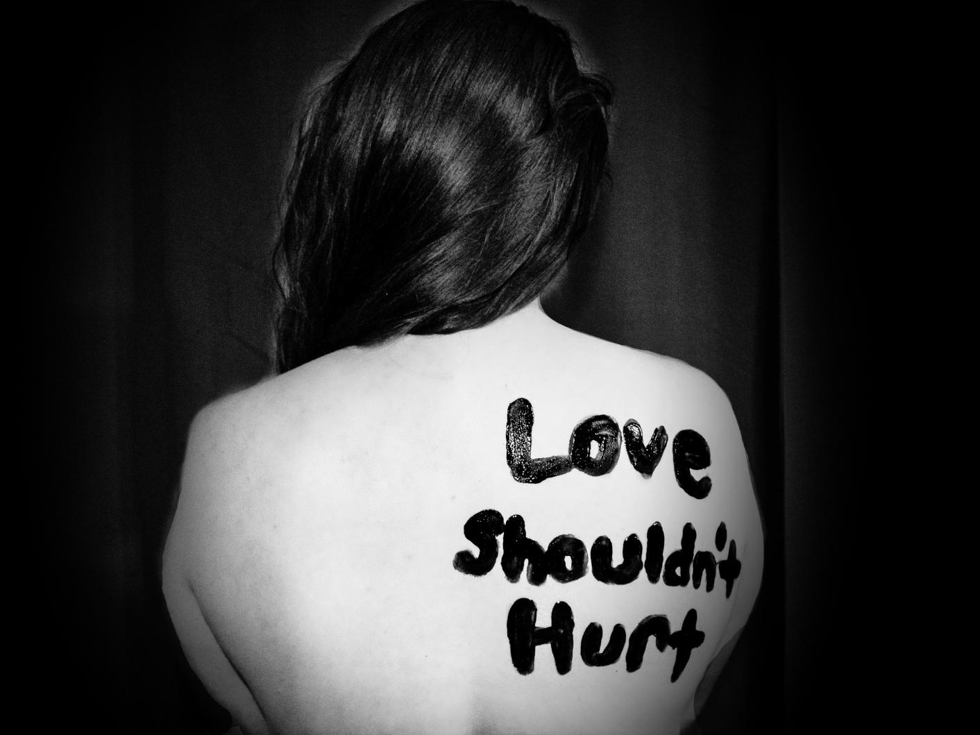 Black and white photo of a women with her back turned to the camera with 'Love shouldn't hurt' painted on her back